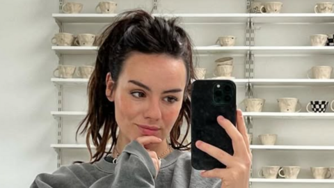 A young woman with dark hair, pulled back into a high ponytail, wearing a grey sweatshirt takes a mirror selfie, behind her is shelving filled with handmade neutral mugs.