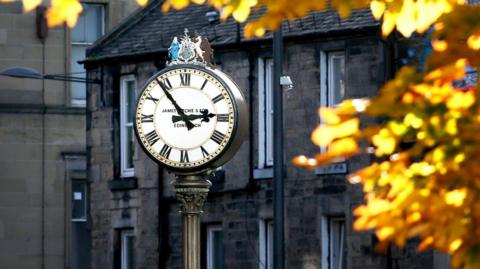 A white clock gace with a coat of arms on top of a black iron pole. It is surrounded by golden leaves and dark-coloured buildings are in the background.