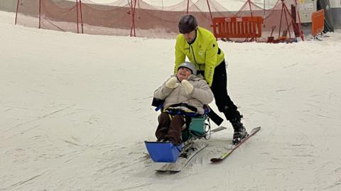 A woman on a sit-ski - an adapted ski with a seat - is guided down a snow slope by a male guide on skis. The woman wears a grey winter coat, white gloves and a grey helmet. The guide wears a hi-vis yellow jacket and black helmet. Red netting can be seen in the background.
