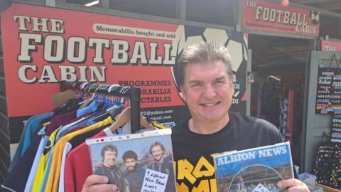 A man holding two football programmes is standing outside the premises of The Football Cabin. There are two signs with its name and football shirts are hung up on the rail outside.