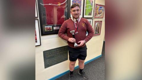 Keenan Pearson in his Netherhall School uniform and holding his trophy while stood in one of the school's corridors. 