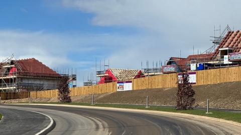 A view of a building site with a wooden fence surrounding the perimeter. Over the fence houses are being built with scaffolding up around them. The sweep of the road at the forefront appears new, but slightly dirty with soil and brick dust. The banks leading up to the fencing are unplanted soil.