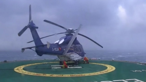 The blue and white helicopter is slightly tilted to one side on a green and yellow helipad. One of the rotor blades is broken. In the background is a stormy sea and clouds.