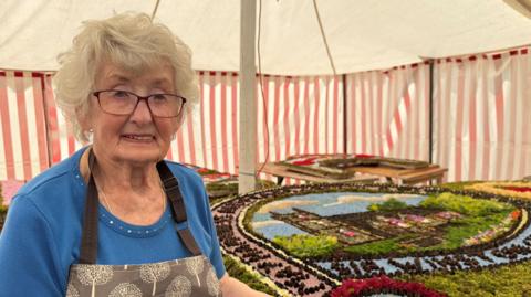 Barbara Hall, a woman wearing a blue top and an apron, is stood next to a colourful well dressing inside a tent.