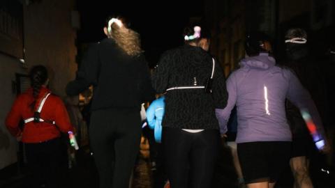 A group of women running in a group in the dark. They are wearing jackets which have hi-vis properties and lights.