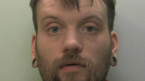 The image shows a close up of a mug shot of a man. He is staring at the camera. 