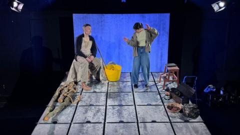 A small stage setup with a man and a woman positioned on a raised, grid‑patterned platform made of square, metal‑looking panels. The lighting is focused on the stage, leaving the surrounding area darker. The platform consists of roughly four rows of large square panels with a textured, slightly worn surface. The man is wearing loose, beige clothing and light tan shoes. A woman stands to the right, mid‑gesture, with one arm raised and the other extended outward. She is wearing wide‑leg trousers, layered clothing including a top and outer garment, and light footwear.