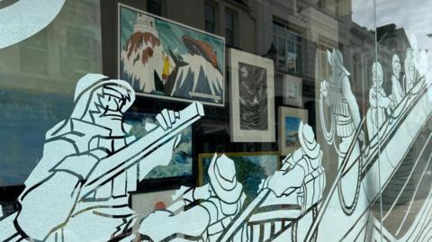 A window display showing an RNLI crew rowing a rescue boat at sea with artwork in the shop inside