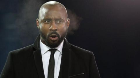 Jason Roberts in a black overcoat and tie exhales on a cold night