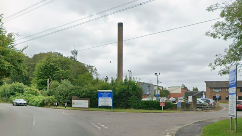 A road with a hospital to the right, showing a large brick chimney, several buildings and a number of signs at the entrance. The road bends to the left and also another one leads to the hospital. There are trees to the right and a car on the road. 