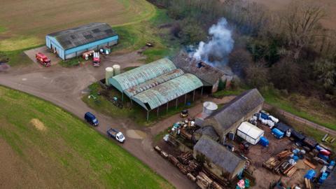 An aerial view of a rural farm showing several large sheds with corrugated roofs. One barn has a hole in the roof with white smoke rising out of it.