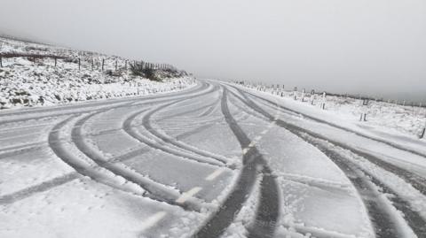 Snowy conditions on the Mountain Road at the Hailwood's Rise. Fence posts and road markers line the snow-covered road which has the tyre tracks of vehicles on it. There is snow cover on the grass at either side.