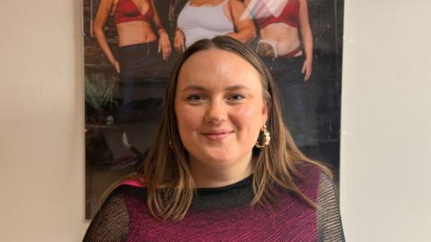 Ellie Brown with long brown hair and gold earrings wearing a black see through top with a hot pink vest underneath. She stands under a poster with three models wearing Recondition clothing. She is smiling.