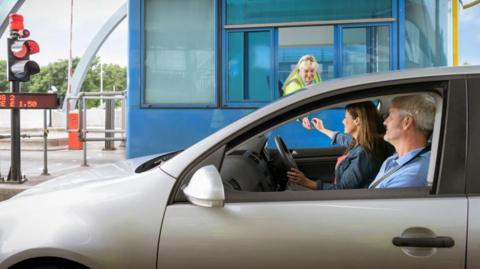 A silver car containing a middle-aged man and woman, stopped at a toll booth.  