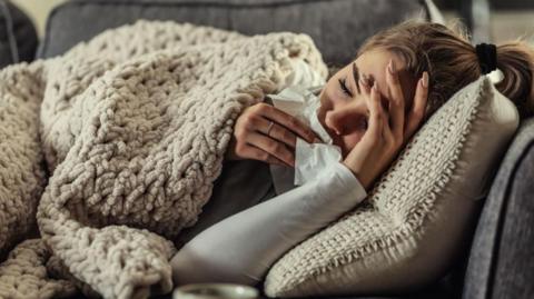 Woman lies on a sofa looking unwell, with a knitted blanket pulled over herself. She holds a tissue in one hand and clutches her forehead, her eyes are closed.