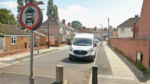 A residential side road with a bungalow and terraced houses. A white van is parked on the road and there is a No Motor Vehicles traffic sign