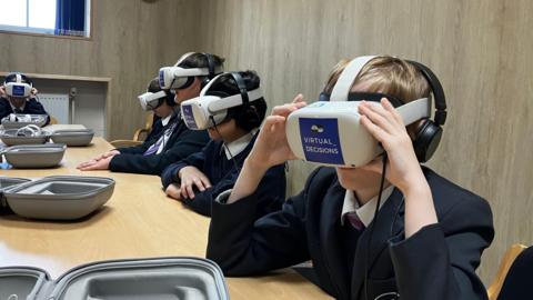 A few children with grey VR headsets on. They have a pair of black headphones on as well a dark navy blue school uniform.