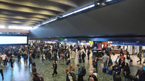 The passenger concourse at Euston station, busy with travellers moving in all directions. A huge screen hangs above running the length of the concourse. The screen is blank. 