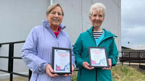 Two women stand together next to a large grey building holding certificates. On the left, Angela Turner has short grey hair and is wearing a purple coat. On the right, Joan Woodhouse has short white hair, and is wearing a green coat.