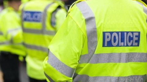 A stock image of police officers wearing yellow high-viz police jackets. The back of an officer's jacket, with the word police is in the foreground. The background is blurred, with other officers, who are all unidentifiable, standing together.