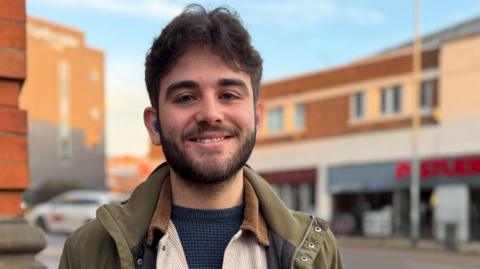Jake Holland, who has dark hair and a beard, is smiling and looking directly at the camera. He is wearing a khaki jacket, cream shirt and grey jumper.