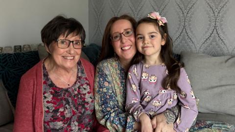 Two woman and a child in the picture. The woman on the left is wearing a red cardigan, a patterned top, has short dark hair and is wearing glasses, she is smiling at the camera. Next to her is a woman in a floral long sleeve top, wearing glasses with shoulder length brown hair and she is smiling. On her knee is a little girl with a long sleeved purple top and long dark hair. They are all sat on a grey sofa.