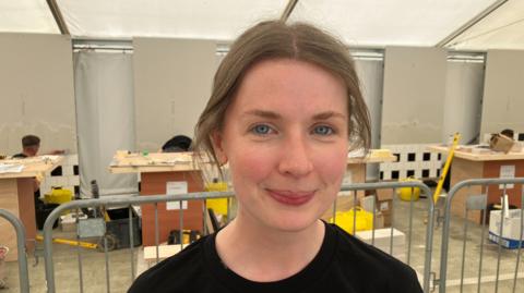 Rebecca Gavigan. She has brown hair tied back, blue eyes, wearing a black t shirt.