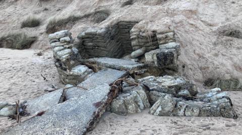 A grey concrete structure is poking out of the sand dunes. Sand bags filled with concrete create the walls of a hut-shaped structure. There are concrete slabs that appear to have collapsed in front of it. The structure is dotted with white splatters which may be lichens or as a result of weather damage.