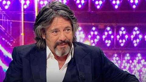 Laurence Llewelyn-Bowen smiling in a smart suit in front of a brightly lit background of pink and purple lights