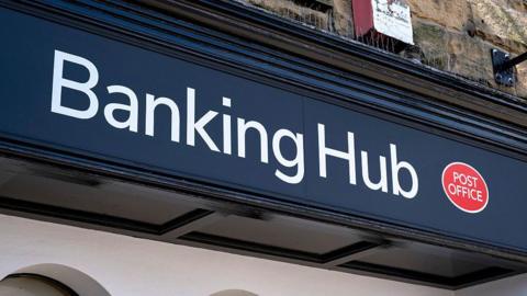 A close-up shot of a blue banking hub sign. The words "Banking Hub" are written in white, with a red Post Office logo next to them.