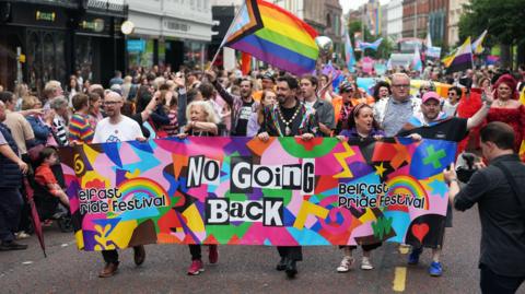 A crowd of people are marching through a city centre. A banner says no going back, Belfast Pride festival. Several people are waving flags.