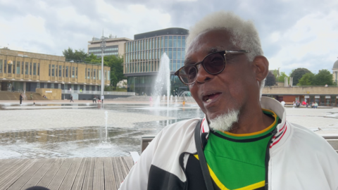 A man with white hair and a beard with sunglasses speaking outside in front of the mirror pool in Bradford