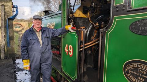 An older man wearing a blue cap, yellow glovas and overalls is standing next to a green steam train.