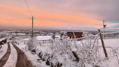 Snow in Mow Cop. A road with snow along it with tyre tracks on it. There is also snow on bushes and in fences. The sky has an orange glow. 