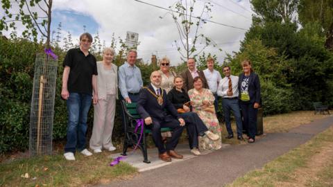 Dignitaries and family members are sat or gathered around a memorial bench. There are sapling trees either side of the bench. The group is looking at the camera, posing for the photo.