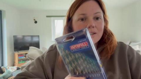 Megan Penny, who has red hair and is wearing a grey hoodie, is sat down showing the camera a packet of drill bits which she received instead of the phone she ordered. 