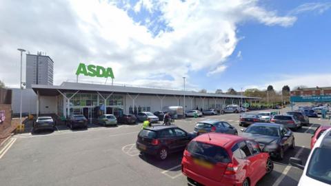 The front of an Asda supermarket - showing the shop, with an Asda sign on the top, and the car park with cars in  