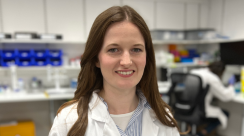 Sarah-Ann Ferguson is stood in the middle of the image smiling. She has long brown hair and is wearing a white lab coat and is standing in a lab.