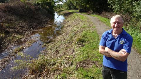 Howard Wilson is standing on a footpath next to a canal. He is crossing his arms and smiling. He is wearing a blue shirt and black jeans.