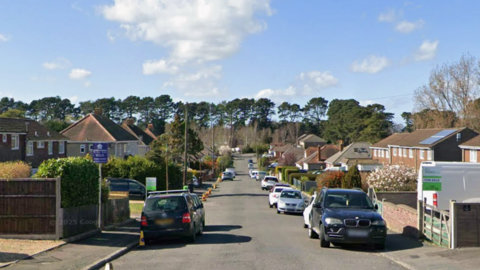 A street with cars and houses either side of a road which stretches down towards some trees.