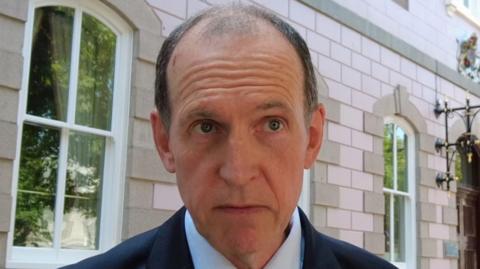 Deputy Steve Ahier, a man with brown/grey hair, wearing a navy blue suit and light shirt. He is standing in front of Jersey's States Assembly building, which has pale pink and granite bricks.