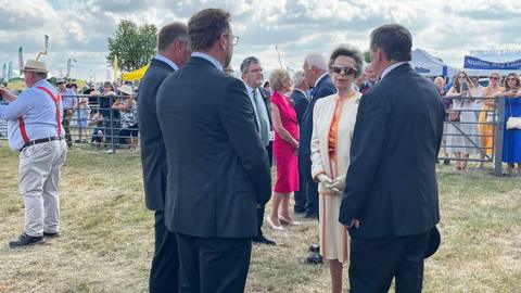 Princess Anne wearing a beige jacket and skirt talks to people at the show with stalls in the background.