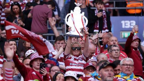 Wigan Warriors fans celebrate their 2024 Challenge Cup final win, one supporter holding a fake cup