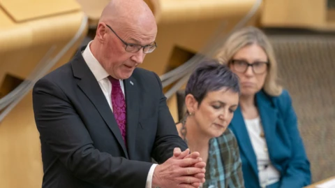 John Swinney has his hands out in front of him as he speaks in the Holyrood chamber. He has a bald head and is wearing glasses. He is wearing a dark suit, white shirt and purple tie