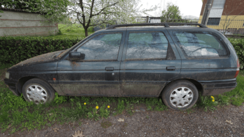 A black estate car with moss growing on the sides and bonnet is parked in  long grass next to a hedge.