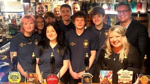 A number of staff are behind the bar in their uniform next to the owners.