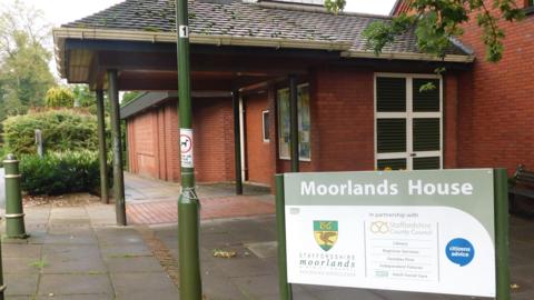 The outside of Moorlands House, a red brick building with a tiled roof shelter near the front door. There is a green and white sign that says "Moorlands House", and has the Staffordshire Moorlands  shield on it.