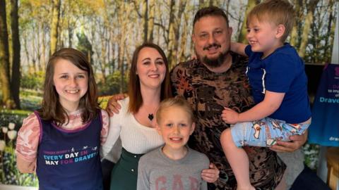 Lynda and Thomas Davies with their three children with woodland wallpaper behind them - all smiling. Lyidan is being carried by his dad Thomas.