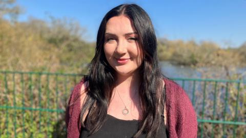 Becca Heritage with black hair stood in front of some trees, bushes and a pond, looking towards the camera. 