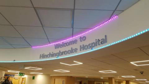 An overhead sign at a hospital that reads "Welcome to Hinchingbrooke Hospital with blue and pink fairy lights underneath and on top